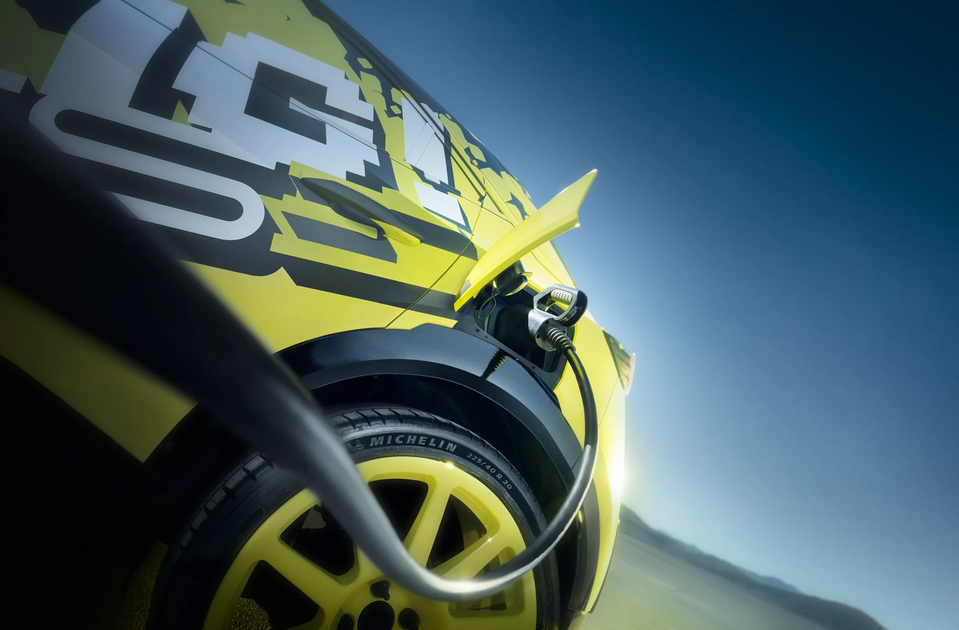 Nahaufnahme eines Elektro-Ladesteckers, der in die Seite eines gelben Opel Rallyeautos gesteckt ist, mit einem Michelin 225/40 R20 Reifen im Vordergrund.