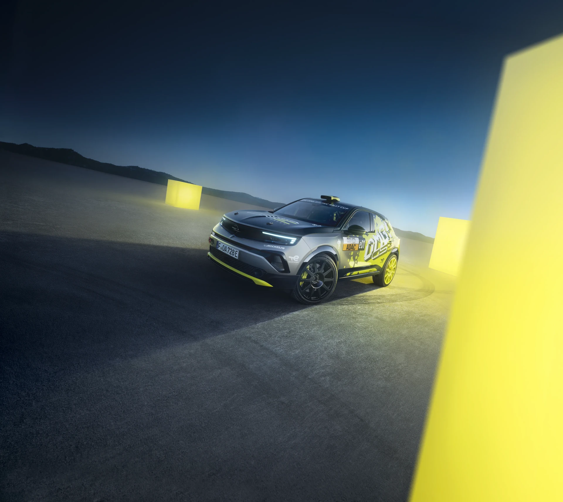 Opel Mokka GSe Elektro-Rallyeauto in grau-gelber Tarnlackierung mit ADAC-Markierungen, in der Abenddämmerung auf einer flachen Fläche umgeben von leuchtend gelben Lichtwürfeln.