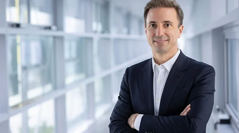 Florian Huettl, CEO von Opel, steht mit verschränkten Armen und lächelnd in einem hellen, modernen Büroflur, gekleidet in einem dunkelblauen Anzug und weißem Hemd.