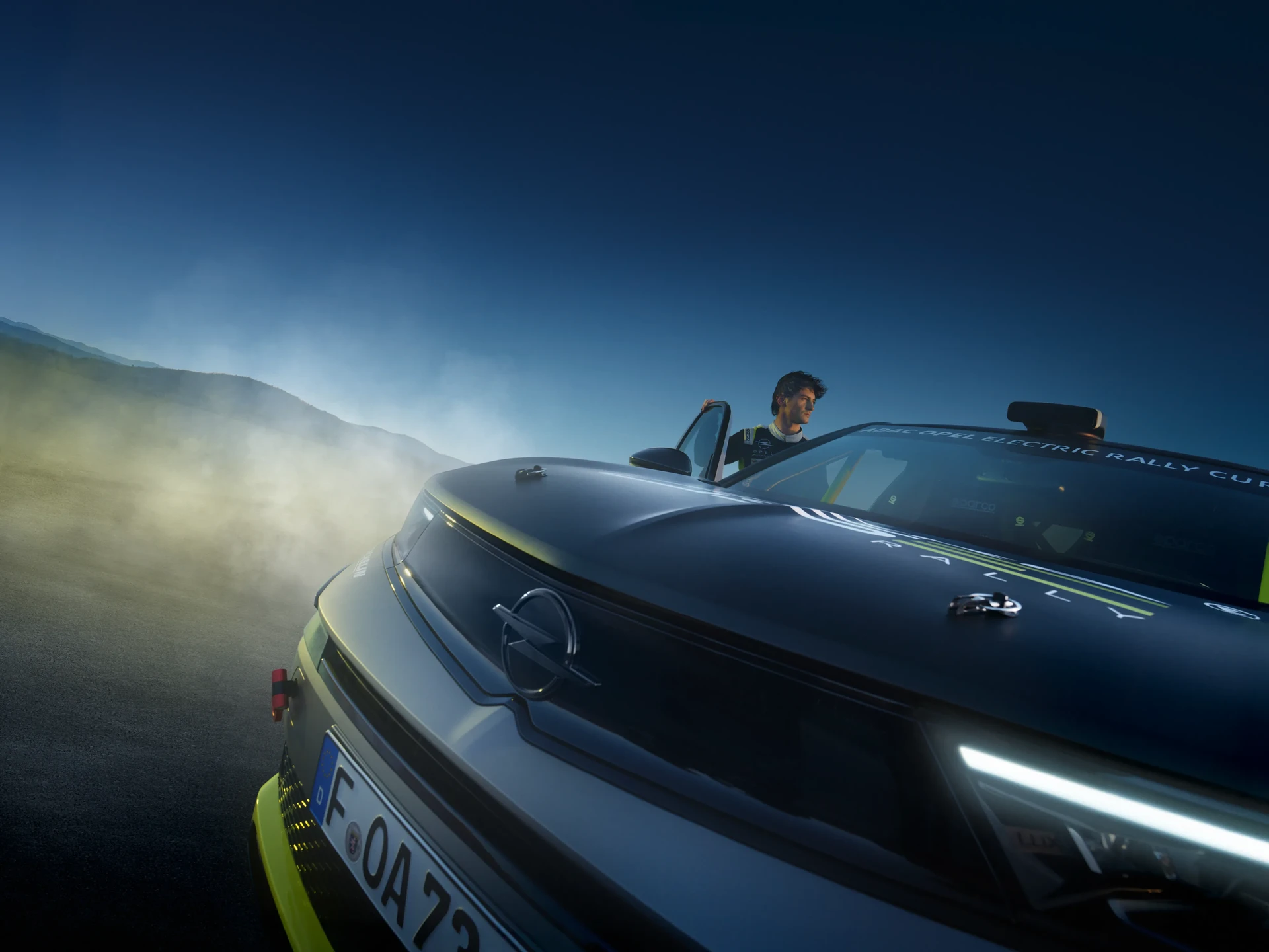 Ein männlicher Fahrer im Rennanzug sitzt in einem Opel Electric Rally Cup-Fahrzeug auf einer dramatisch nebligen Straße in der Abenddämmerung, mit Bergen im Hintergrund. Das dunkel lackierte Rallyefahrzeug weist neongelbe Akzente, das Opel-Logo auf der Motorhaube und helle LED-Scheinwerfer auf.