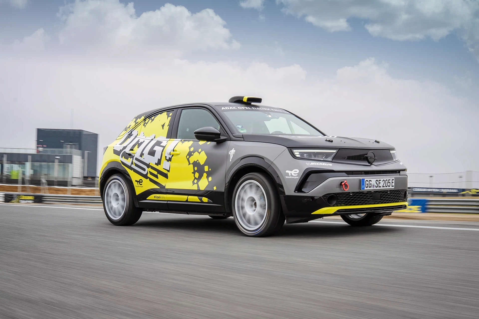 Opel Mokka-e Elektro-Rallyeauto in schwarz-gelber GSe-Rennlackierung mit ADAC Opel Electric Rally Cup- und TotalEnergies-Branding, fahrend auf einer Rennstrecke mit Bewegungsunschärfe im Hintergrund.