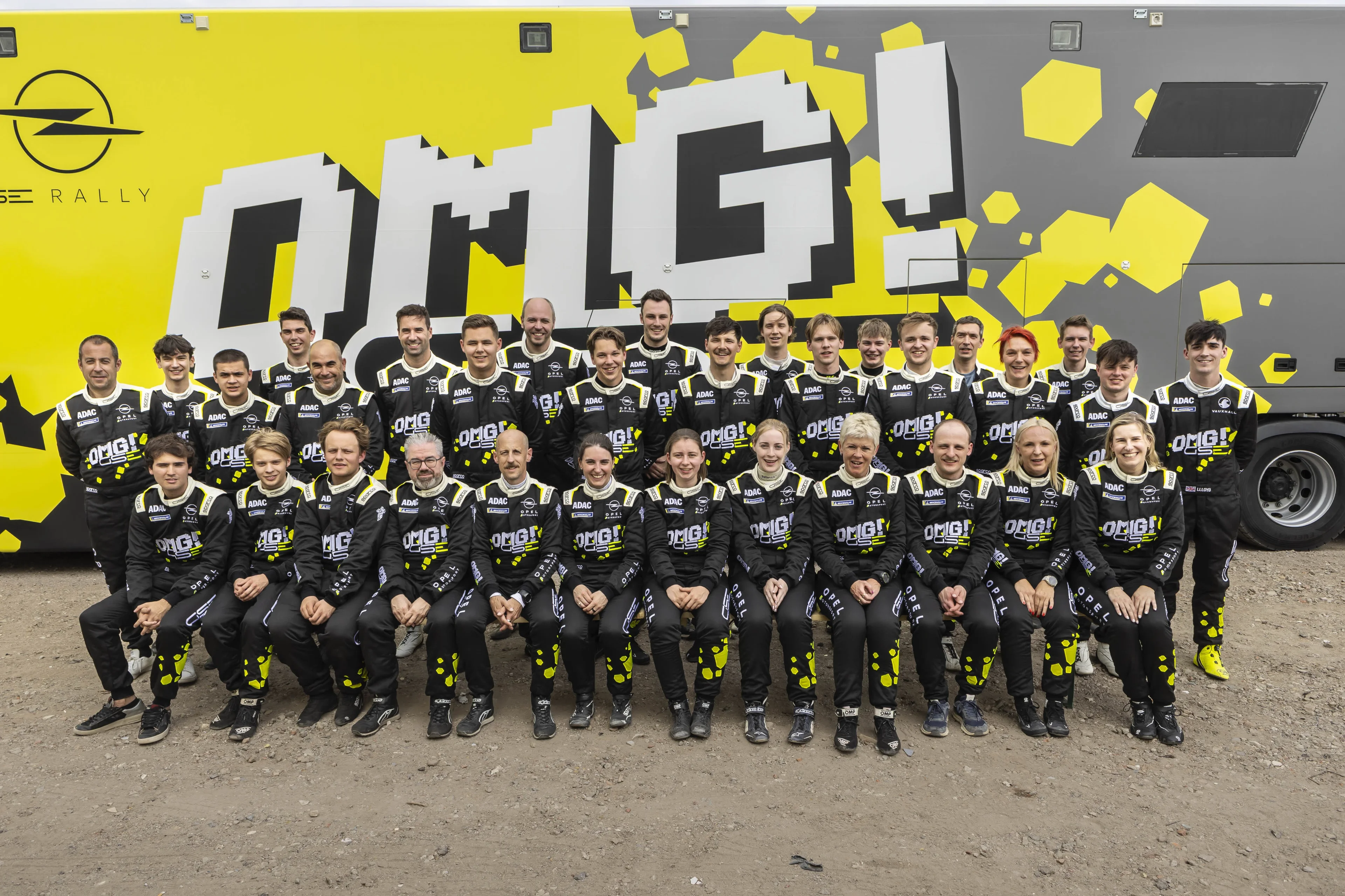 Group photo of around thirty drivers and co-drivers in matching black Opel and ADAC racing suits with "OMG! GSE" graphics, arranged in two rows in front of the Opel GSE Rally team truck in a gravel service park.