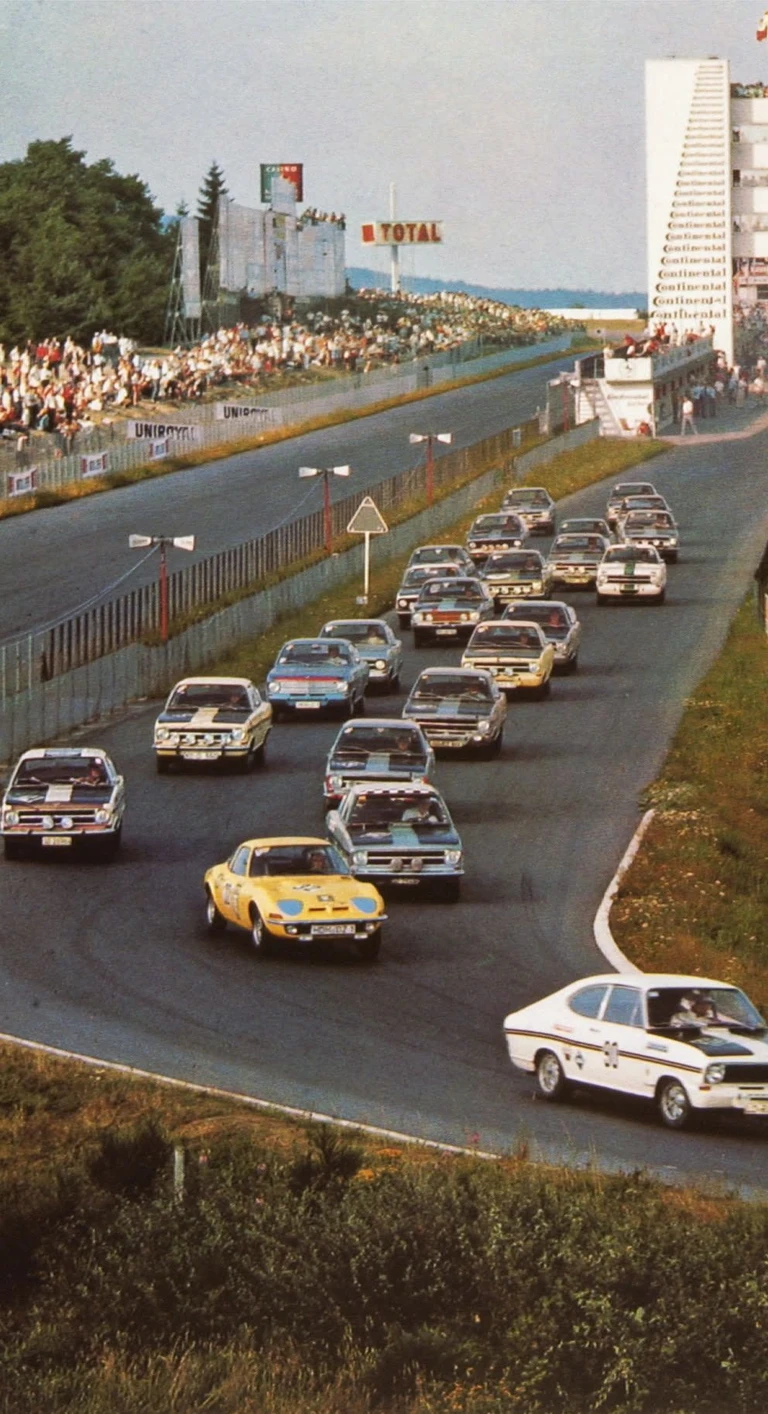 Archive photo of a touring car race start seen from above: a white pace car leads a large field of saloon racing cars down the track past packed grandstands, a tall control tower and a Total advertising board.