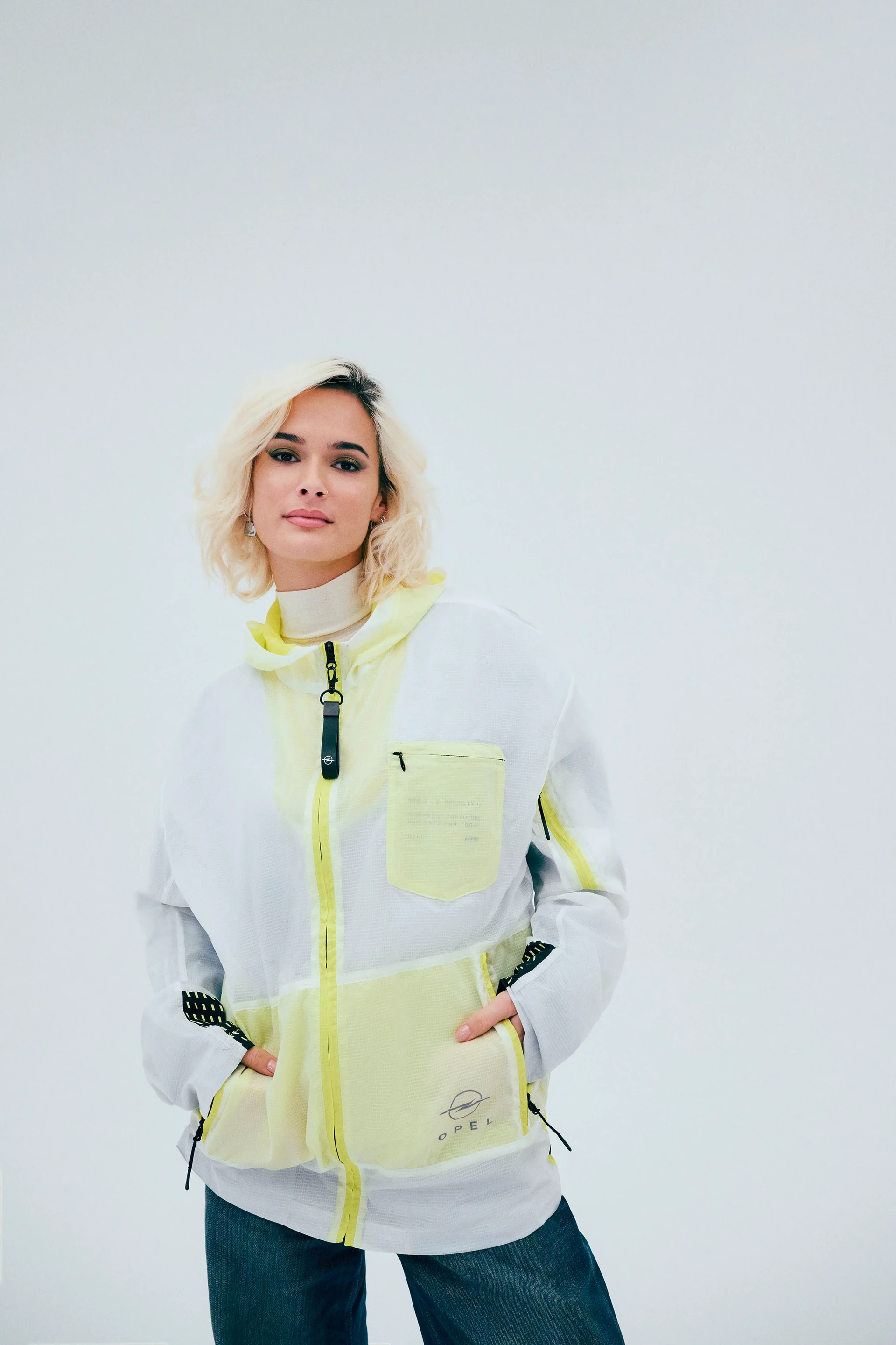 Eine junge Frau mit kurzem blondem Haar trägt eine weiße und neongelbe Opel-Fleecejacke mit Reißverschluss, kombiniert mit einem cremefarbenen Rollkragenpullover und dunkler Jeans, vor einem hellgrauen Hintergrund.