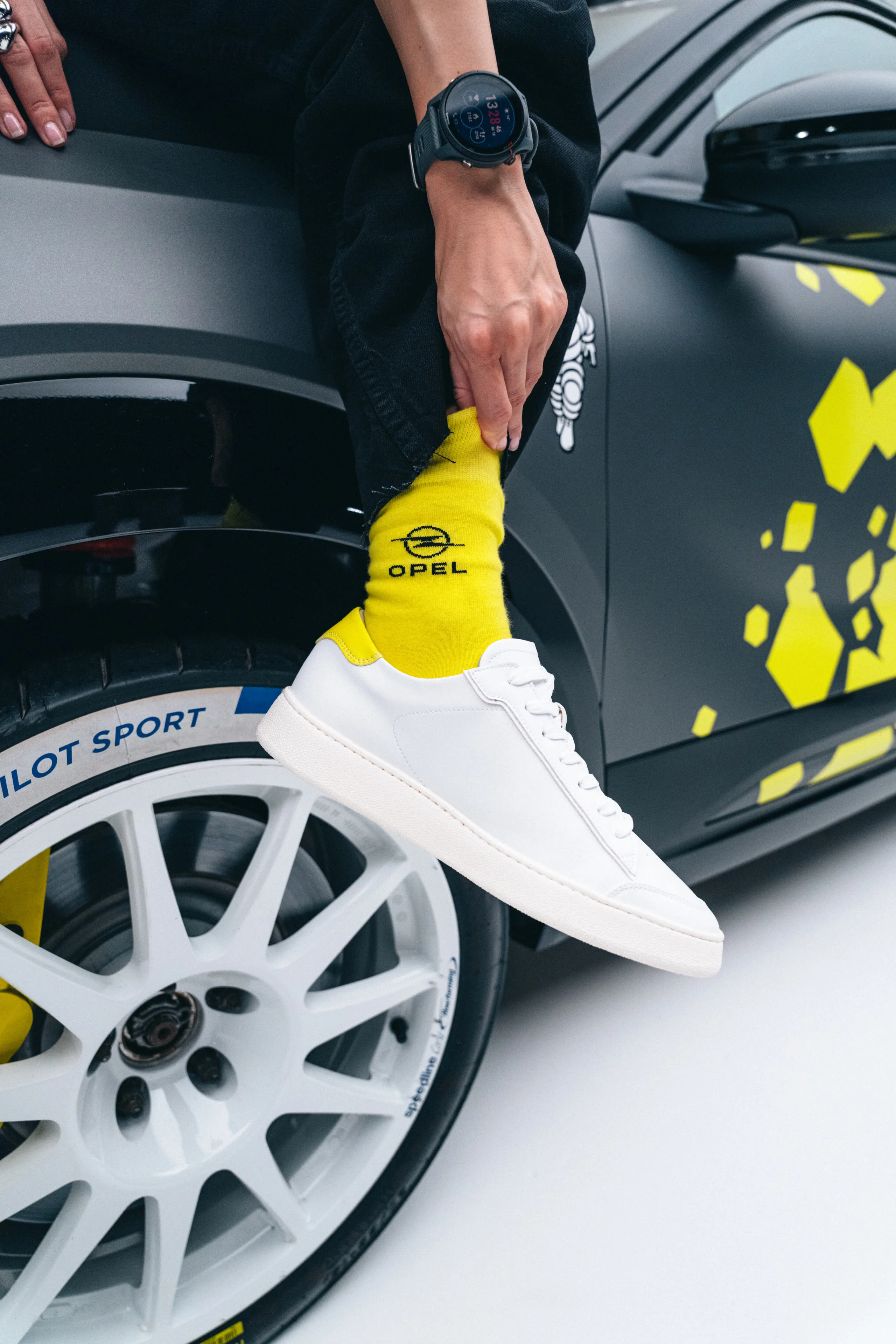 A person's lower leg in yellow Opel-branded socks and white sneakers, resting against the wheel arch of a yellow and black Opel rally car.