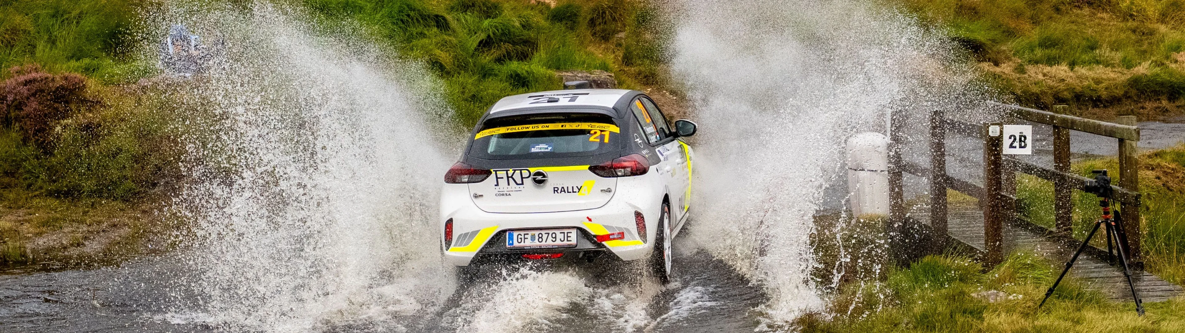 White Opel Corsa Rally car with yellow accents carrying number 21, seen from behind as it powers through a water splash on a moorland stage, throwing spray high on both sides, with spectators and photographers watching from the heather-covered hillside above.