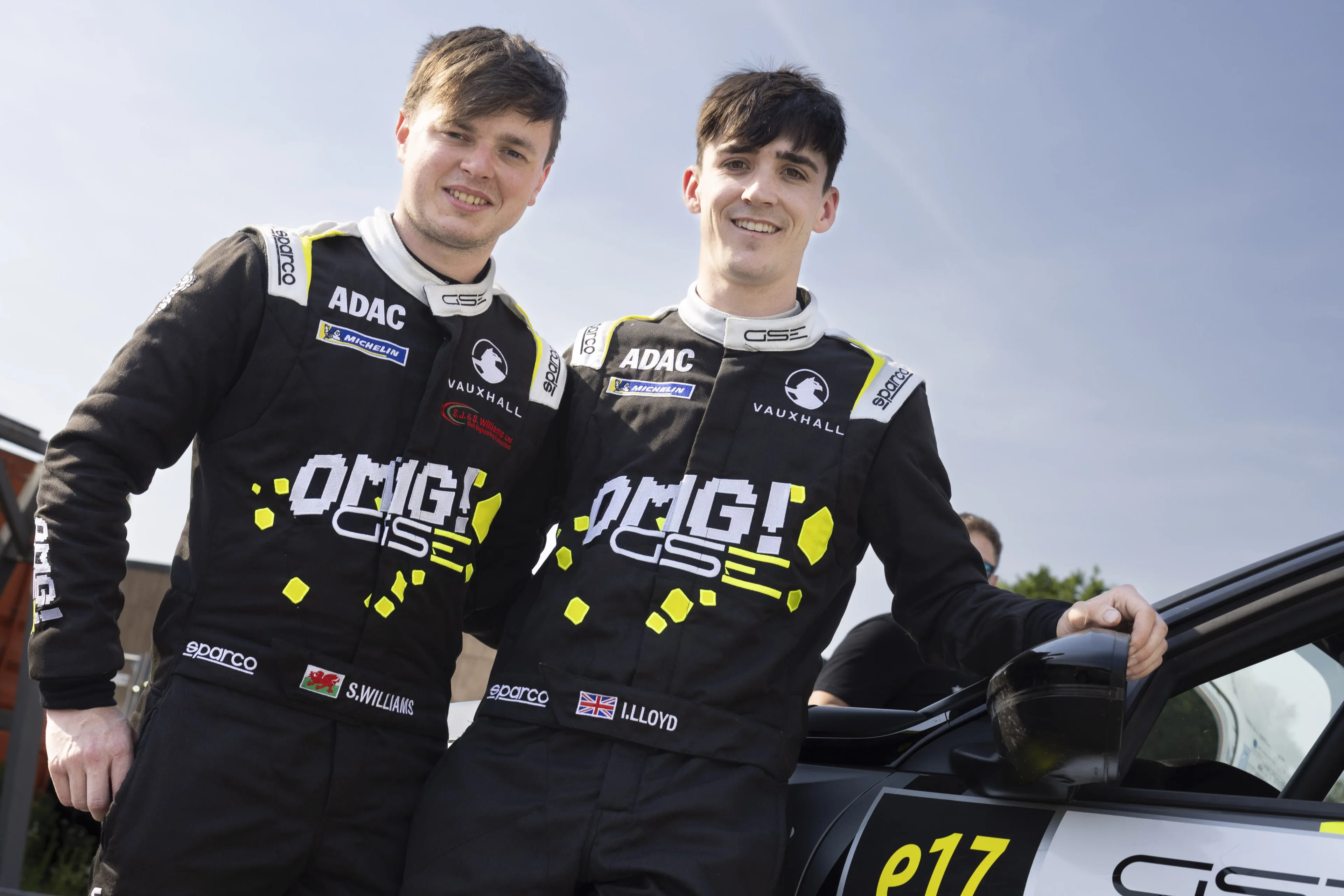Two crew members in black ADAC and Vauxhall racing suits with "OMG! GSE" graphics, standing beside their rally car with the number e17 on the door.