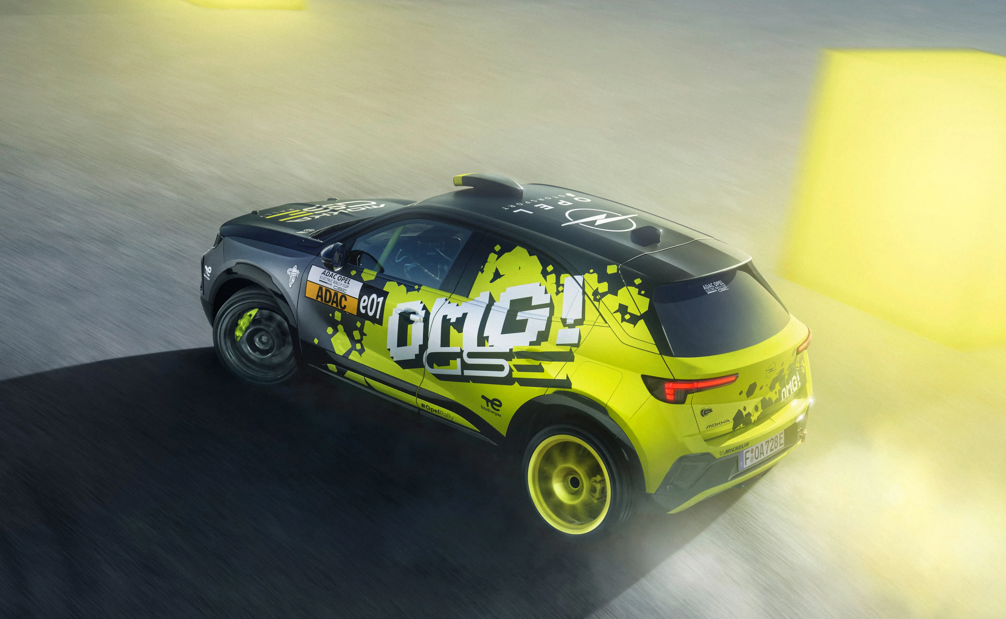Neon yellow and black Opel Mokka GSE Rally car in "OMG! GSE" livery, seen from above and behind while driving, showing the full side graphics, the Opel Motorsport lettering on the roof and the yellow rally wheels.