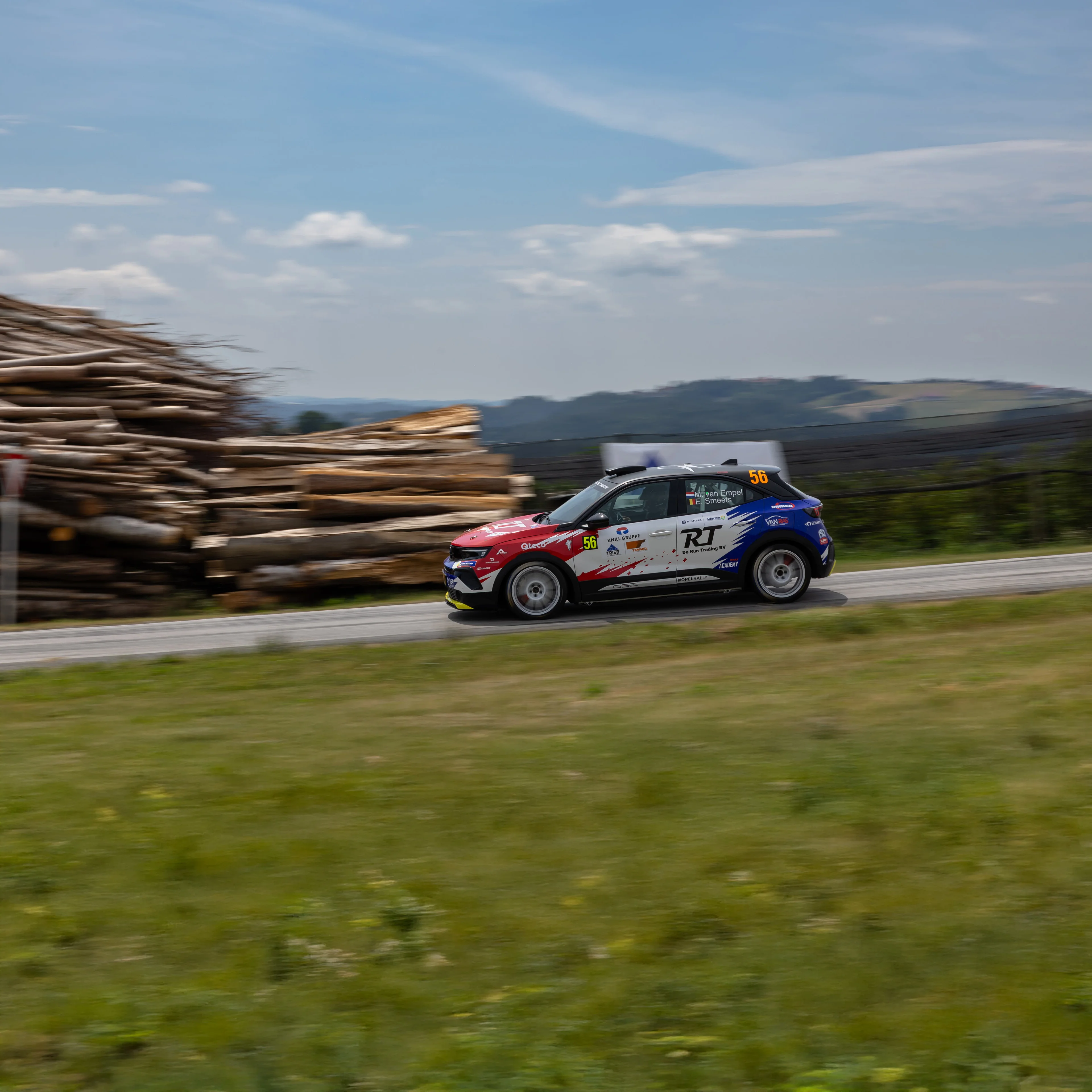 Opel Mokka GSE Rally car in red, white and blue livery carrying number 56, seen in side profile at speed on a tarmac stage past a stack of felled timber, with rolling hills behind and the background blurred by a panning shot.