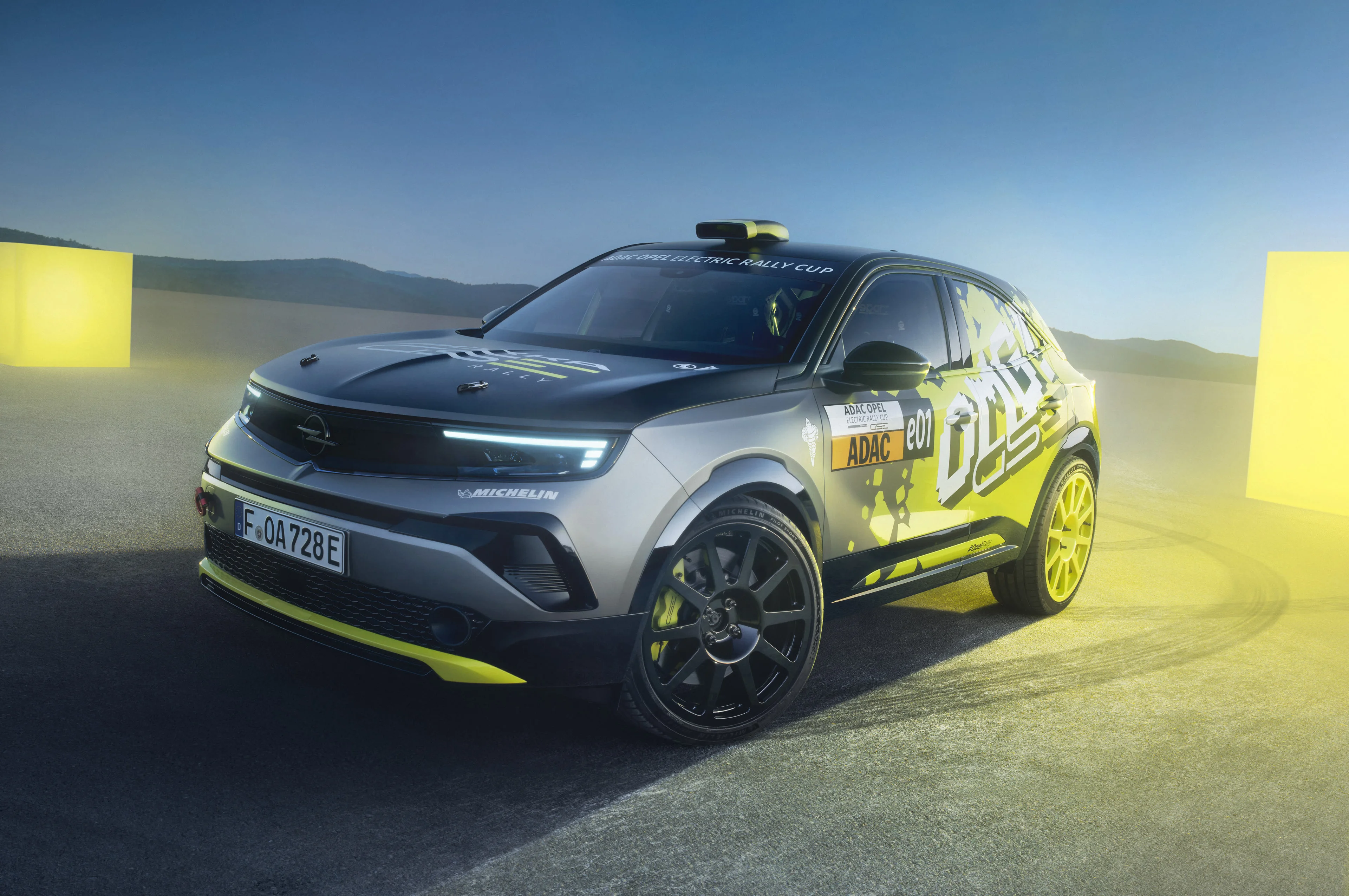 Opel Mokka GSE Rally car in grey with yellow and black "OMG GSE" graphics, standing still on tarmac at dusk between two glowing yellow course markers, seen from the front three-quarter with its daytime running lights on.
