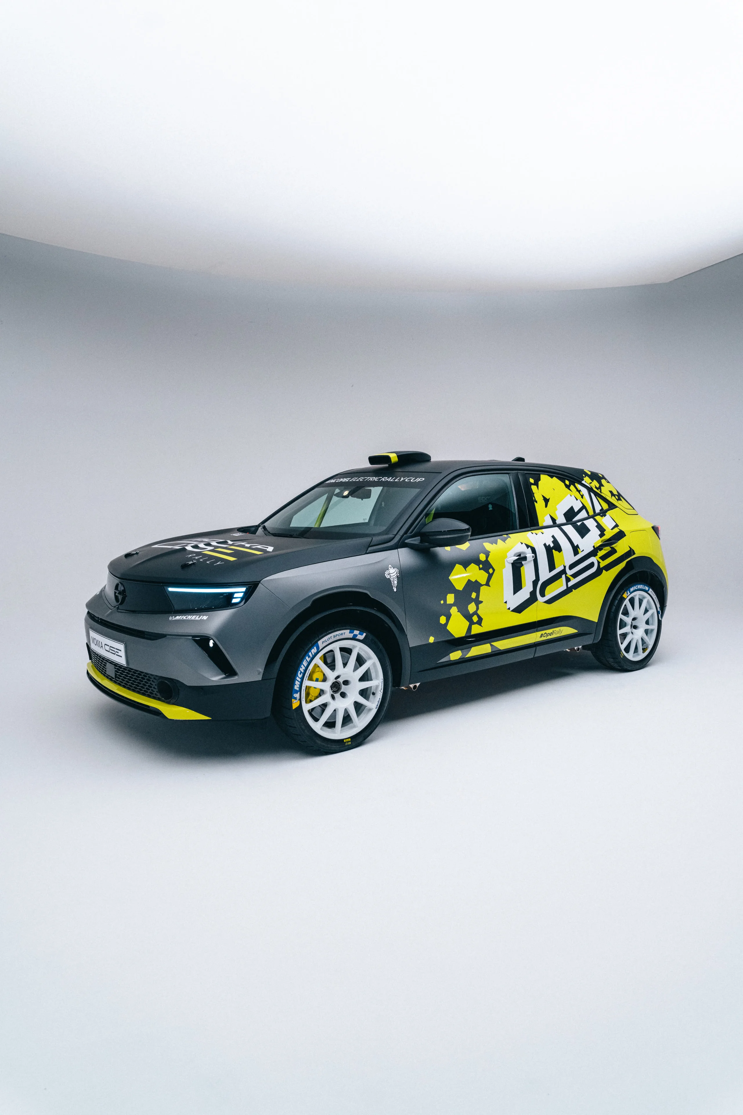 Opel Mokka GSE rally car in grey with yellow and black "OMG GSE" graphics and white rally wheels, photographed stationary in a plain studio setting from the front three-quarter.