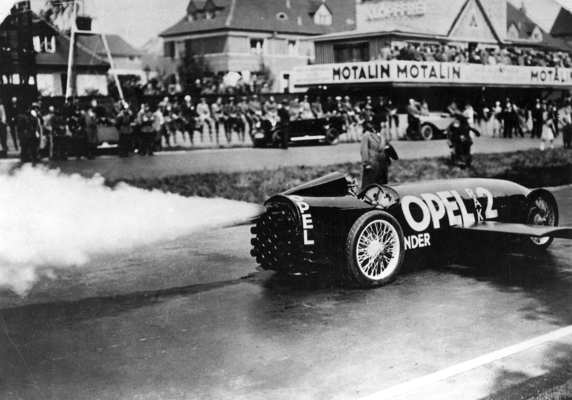 Black-and-white photo of the black Opel RAK 2 rocket car speeding past a crowd and grandstands with "Motalin" banners, white smoke streaming from its rear rockets.