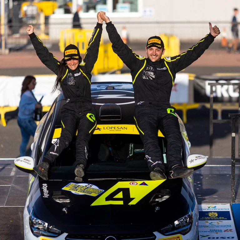 A rally driver and co-driver in black and yellow racing suits sitting on the bonnet of their number 4 car, hands joined and arms raised in celebration.